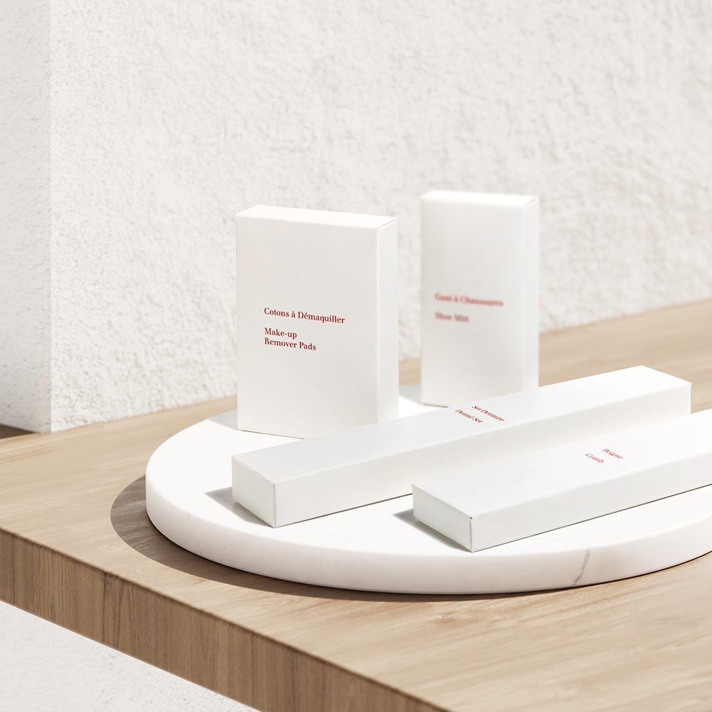 A minimalist display of white packaging for makeup remover pads and skincare products, featuring simple red text labels, arranged on a round marble surface against a textured white wall.