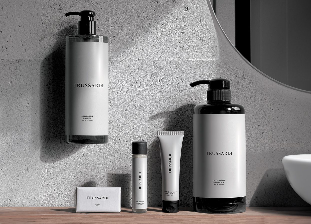 A minimalist arrangement of Trussardi skincare products on a wooden surface against a concrete wall, featuring various sizes of bottles and tubes.