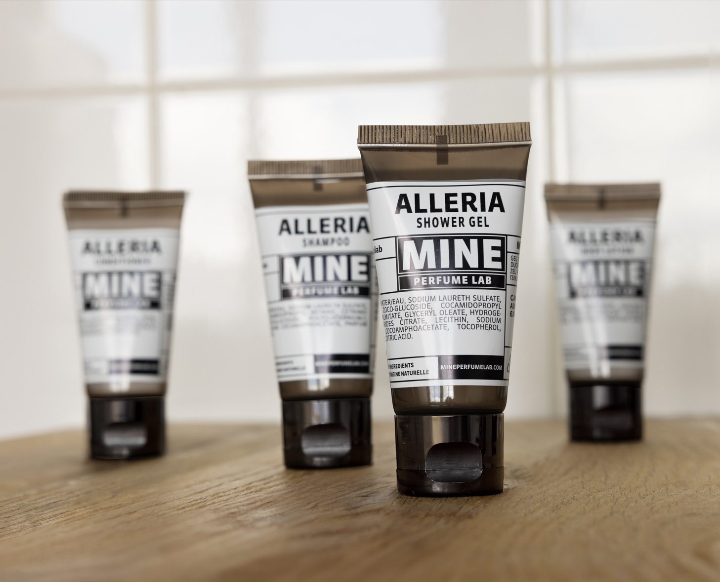 Four tubes of Alleria shower gel and shampoo arranged on a wooden surface, with a clean, modern design and black and white labeling. The background features light-colored tiles, creating a neutral setting.