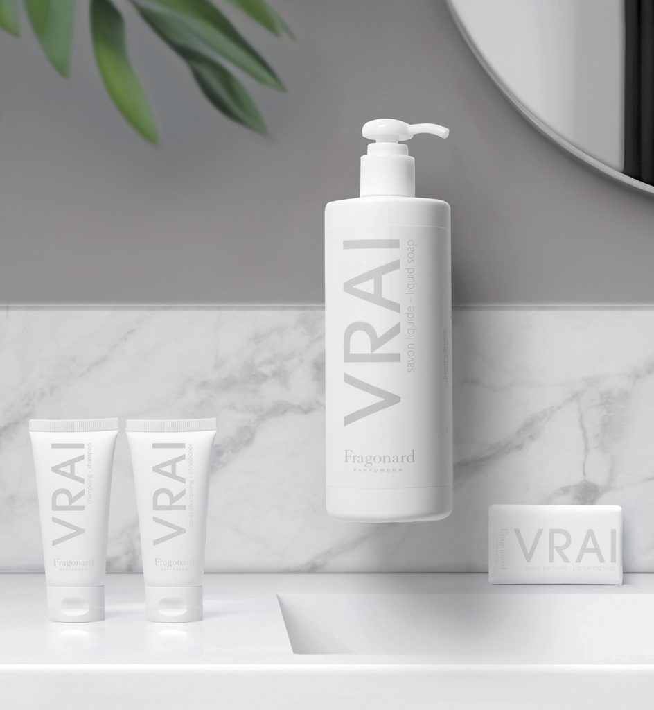 A minimalist bathroom scene featuring various skincare products labeled "VRAI," including a large liquid soap bottle, two small tubes, and a bar soap, set against a marble surface and a circular mirror.