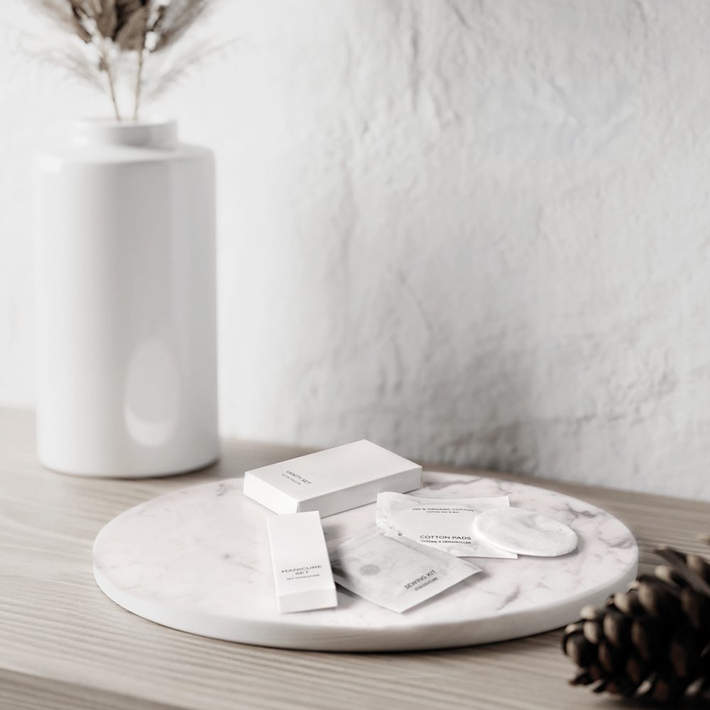 A minimalist display featuring white packaging for skincare products arranged on a round marble tray, alongside a small vase and a pinecone.