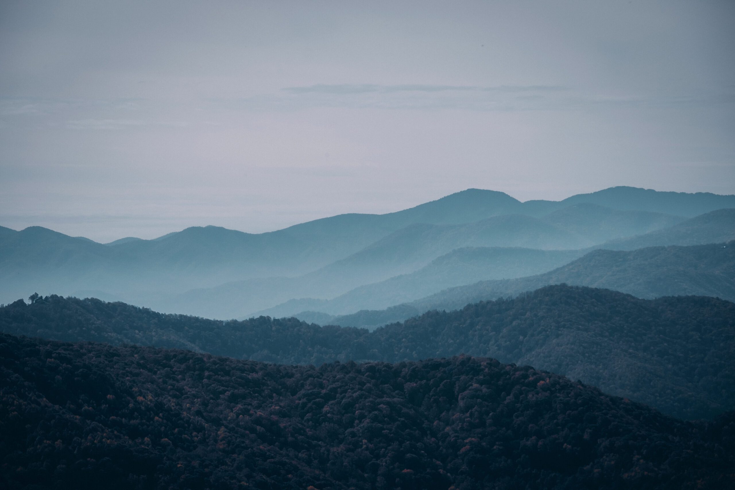 A panoramic view of layered mountains shrouded in a misty haze, creating a serene, atmospheric landscape. The varying shades of blue and green highlight the natural contours of the terrain.