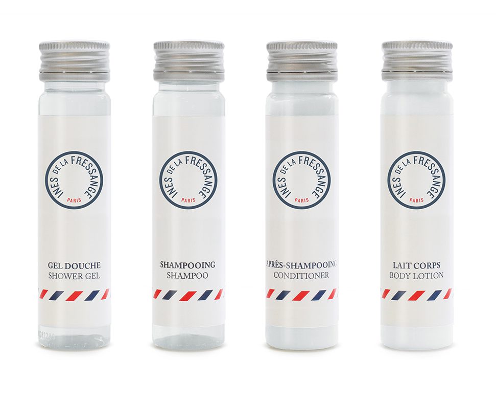 Four transparent bottles lined up, each labeled with white text: "Gel Douche Shower Gel," "Shampooing Shampoo," "Près-Shampooing Conditioner," and "Lait Corps Body Lotion." Each bottle features a simple design with a circular logo.