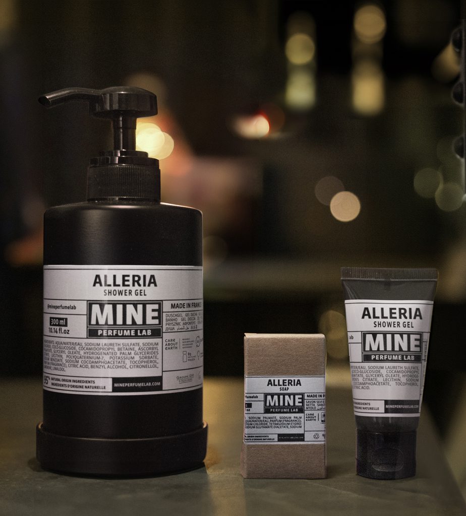 A black pump bottle of Alleria shower gel alongside a small tube and a box, all featuring minimalist labels with the word "MINE" prominently displayed. The background is softly blurred with warm lighting.