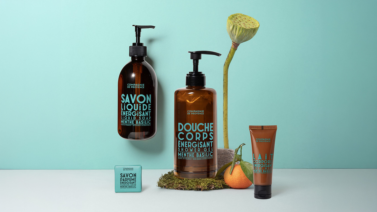 Set of amber bottles and skincare products labeled ‘Compagnie de Provence’ arranged on a turquoise background with a lotus stem and citrus fruit.