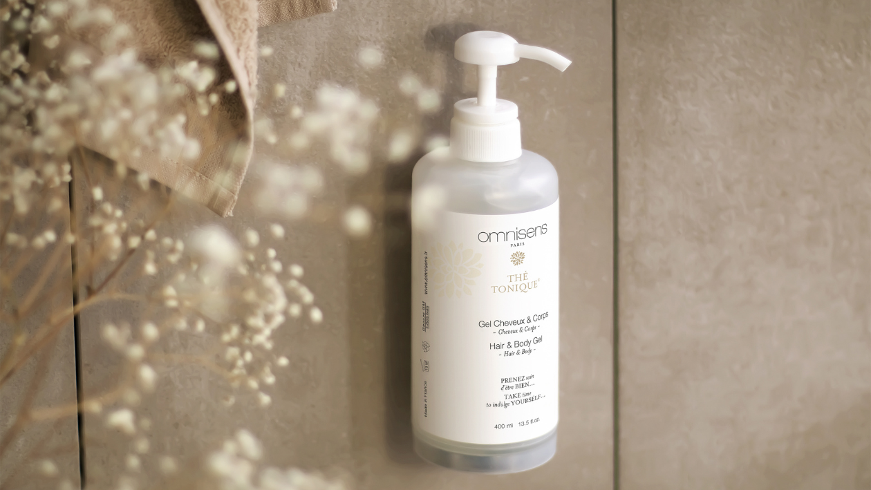 Omnisens Paris “Thé Tonique” hair and body gel bottle mounted on a tiled bathroom wall with soft floral elements in the foreground.