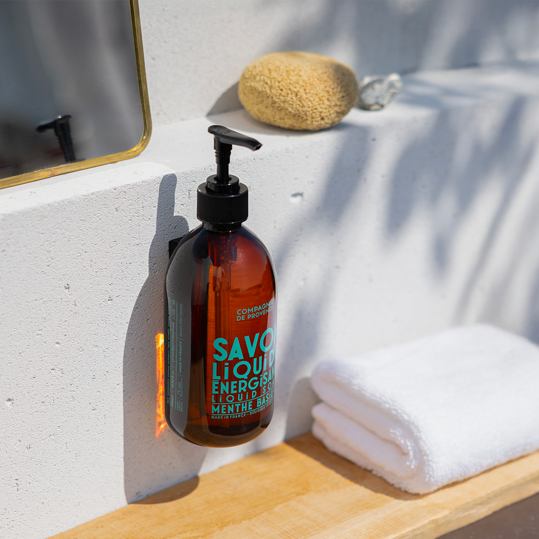 Compagnie de Provence liquid soap bottle with pump mounted by a sink, next to a towel and sponge in a sunlit bathroom setting