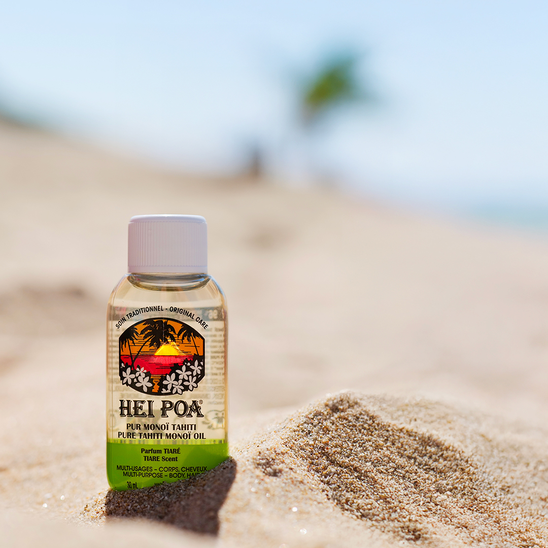 Hei Poa monoi oil bottle placed on sand at the beach with a blurred tropical background