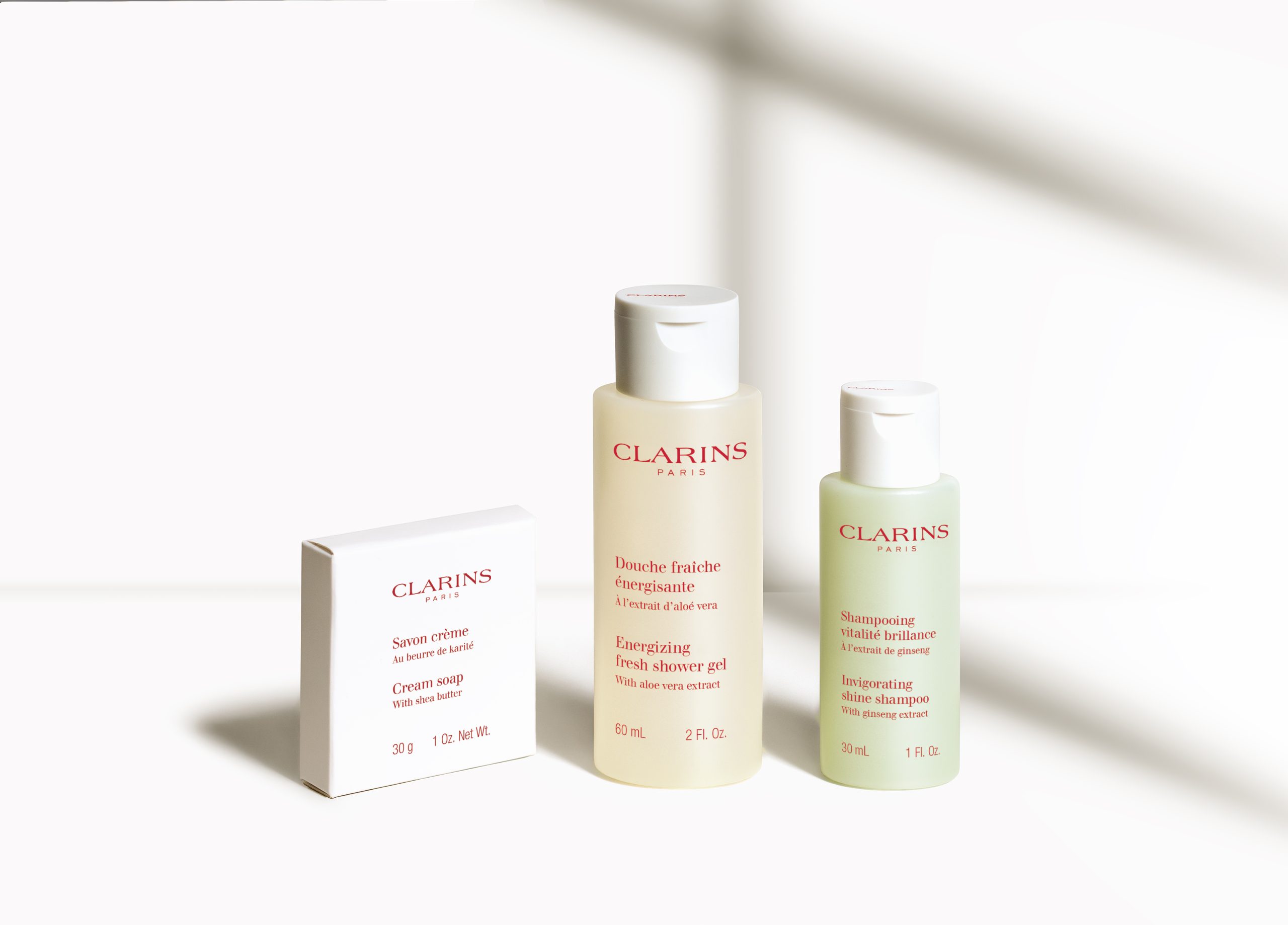 Clarins produtos de higiene pessoal em tamanho de viagem, incluindo sabonete em creme, gel de banho refrescante e champô brilhante, dispostos sobre um fundo branco brilhante.