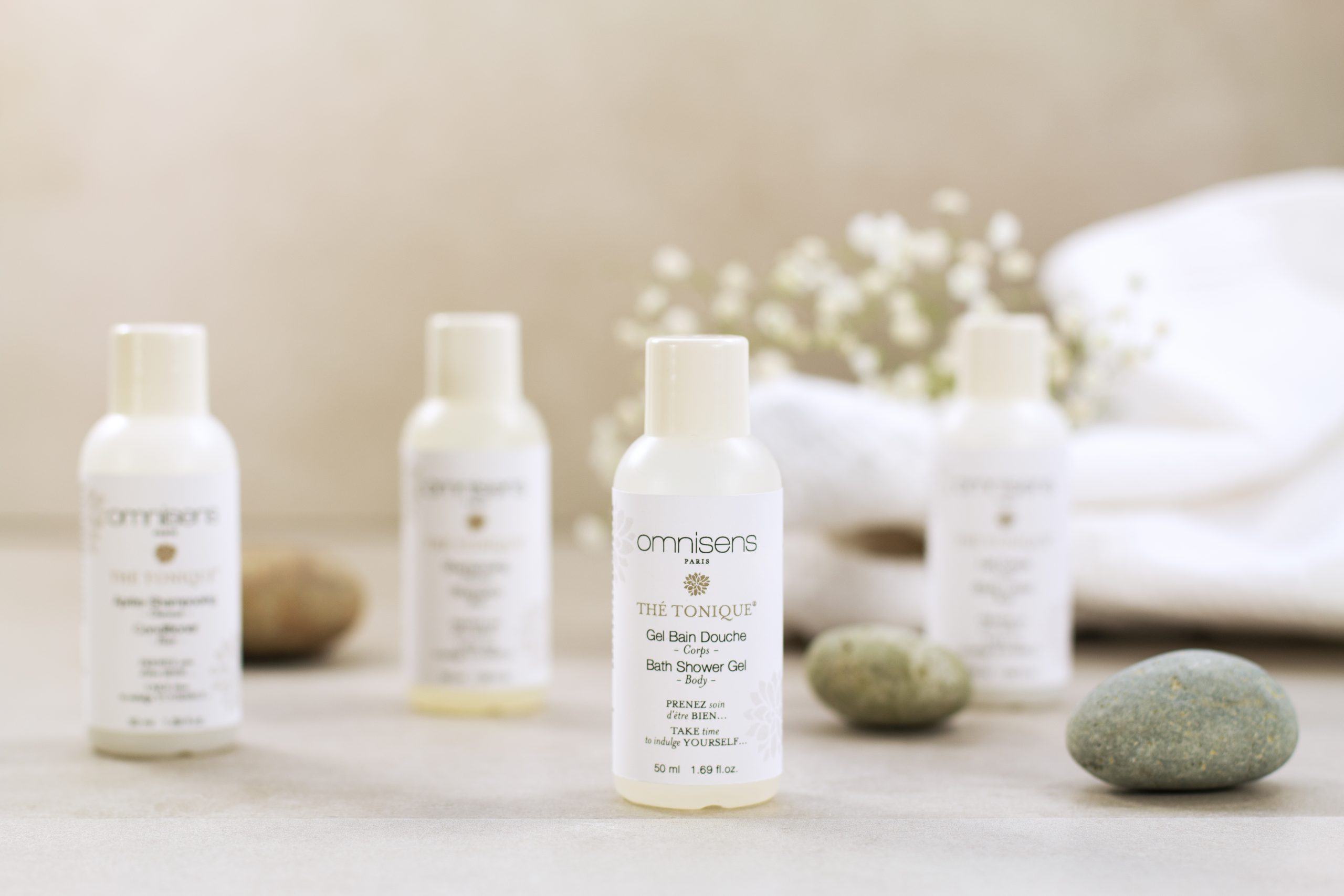 Mini Omnisens Paris “Thé Tonique” bath and shower gel bottle centered among spa-style stones and softly folded towels.