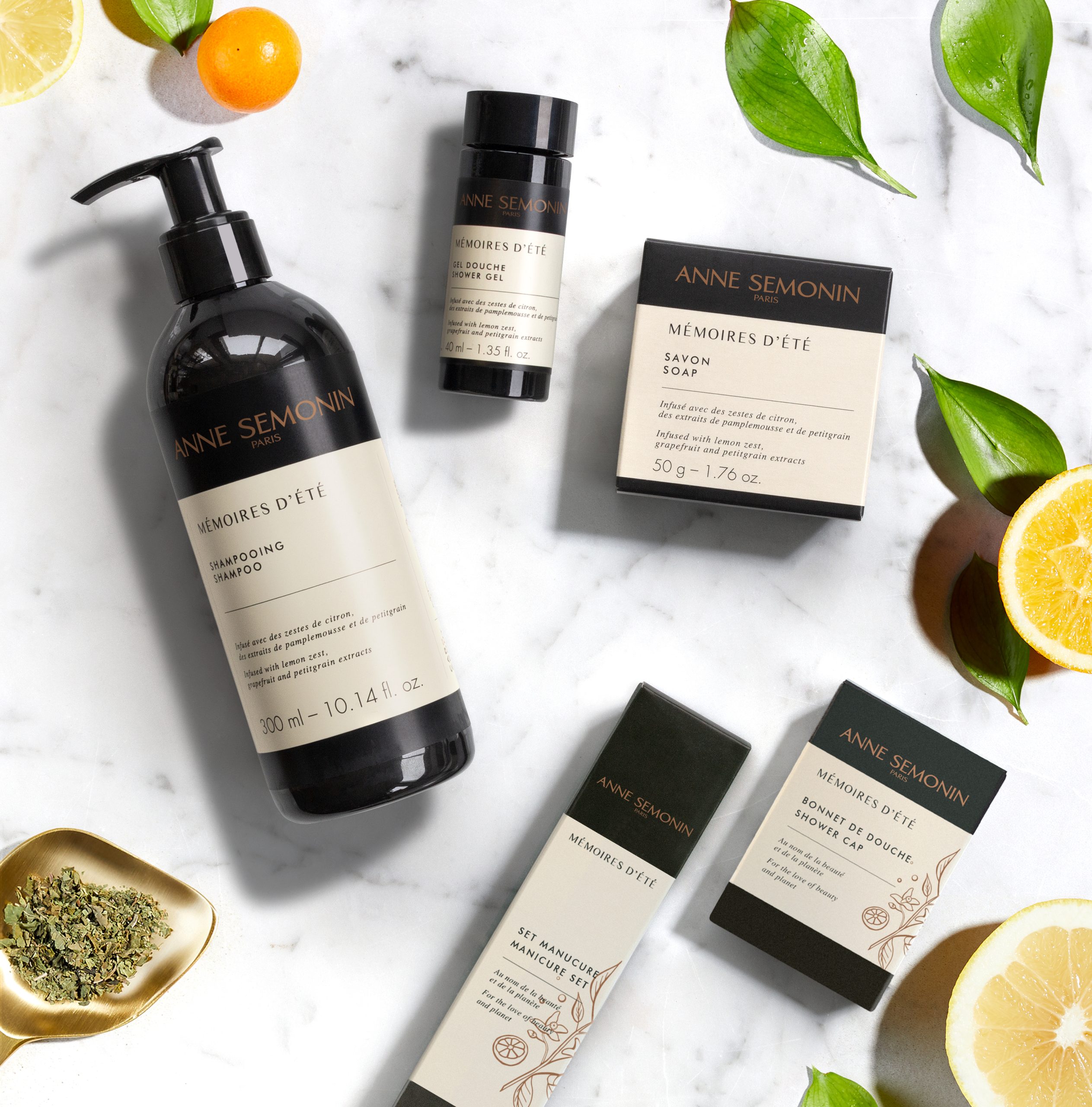 Anne Semonin De shampoo, zeep en toiletartikelen uit de “Mémoires d’Été”-lijn liggen plat op een marmeren oppervlak, omringd door citrusvruchten en groene bladeren.