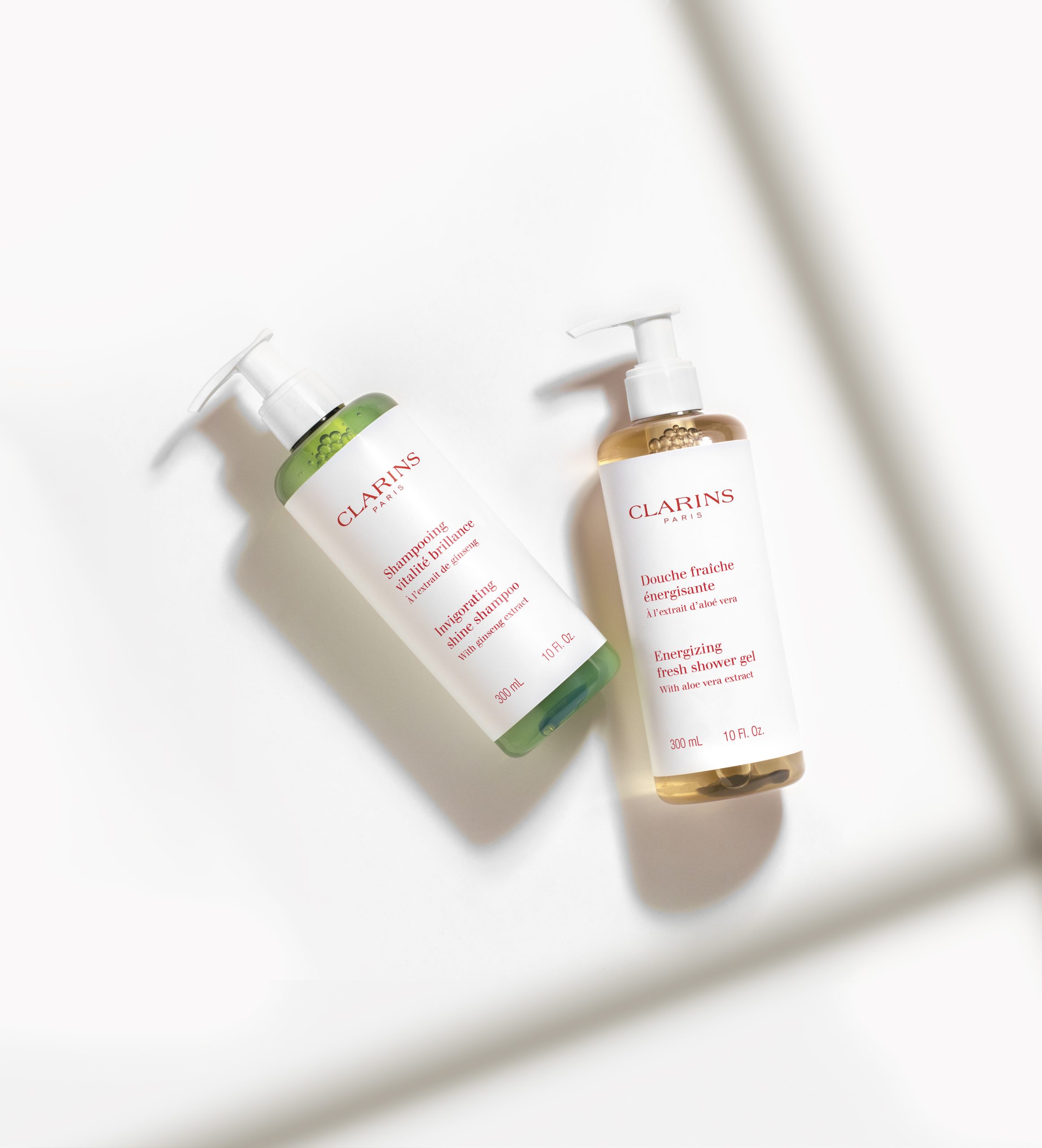 Clarins Frascos de champô revigorante e brilhante e de gel de banho energizante e refrescante, dispostos sobre uma superfície branca com luz natural suave.