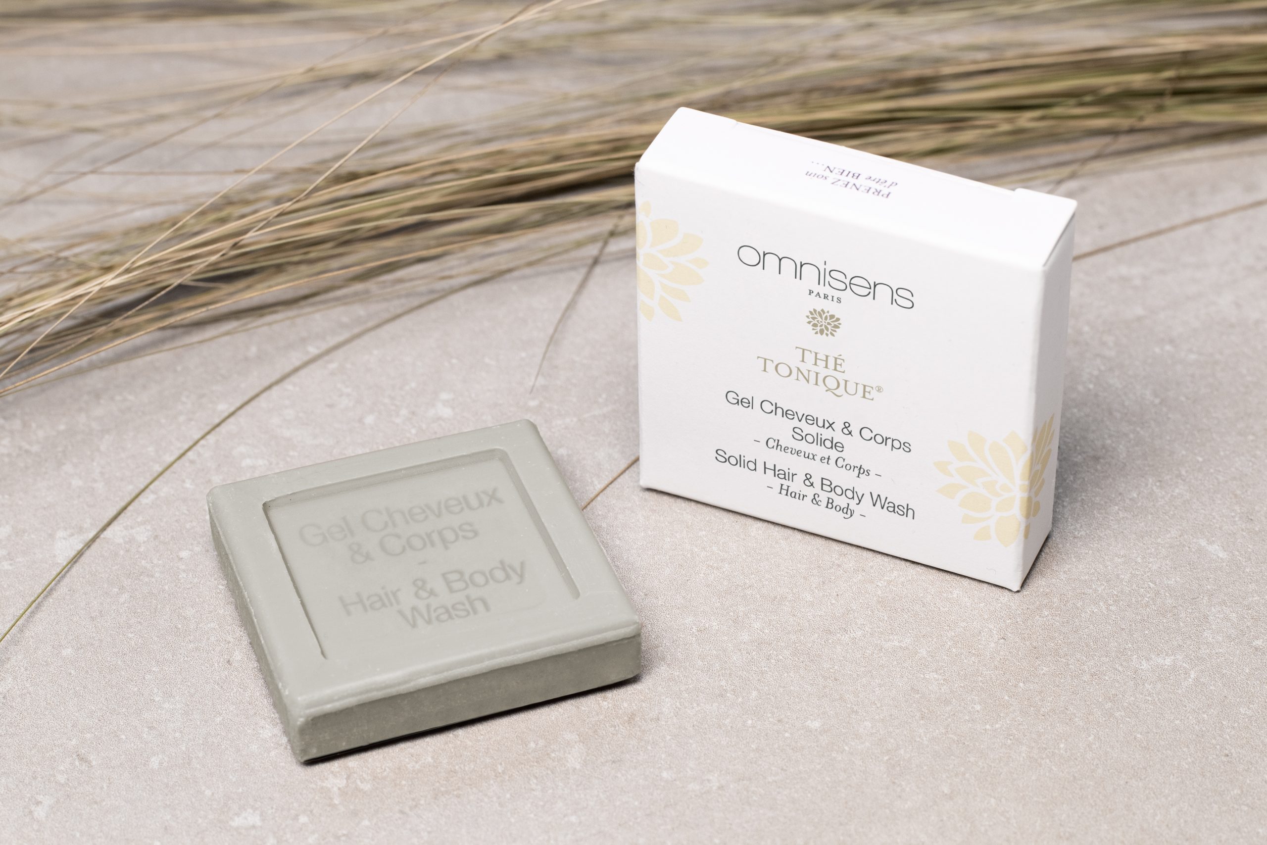 Omnisens Paris “Thé Tonique” solid hair and body wash bar placed beside its packaging on a light stone surface with dried grasses.