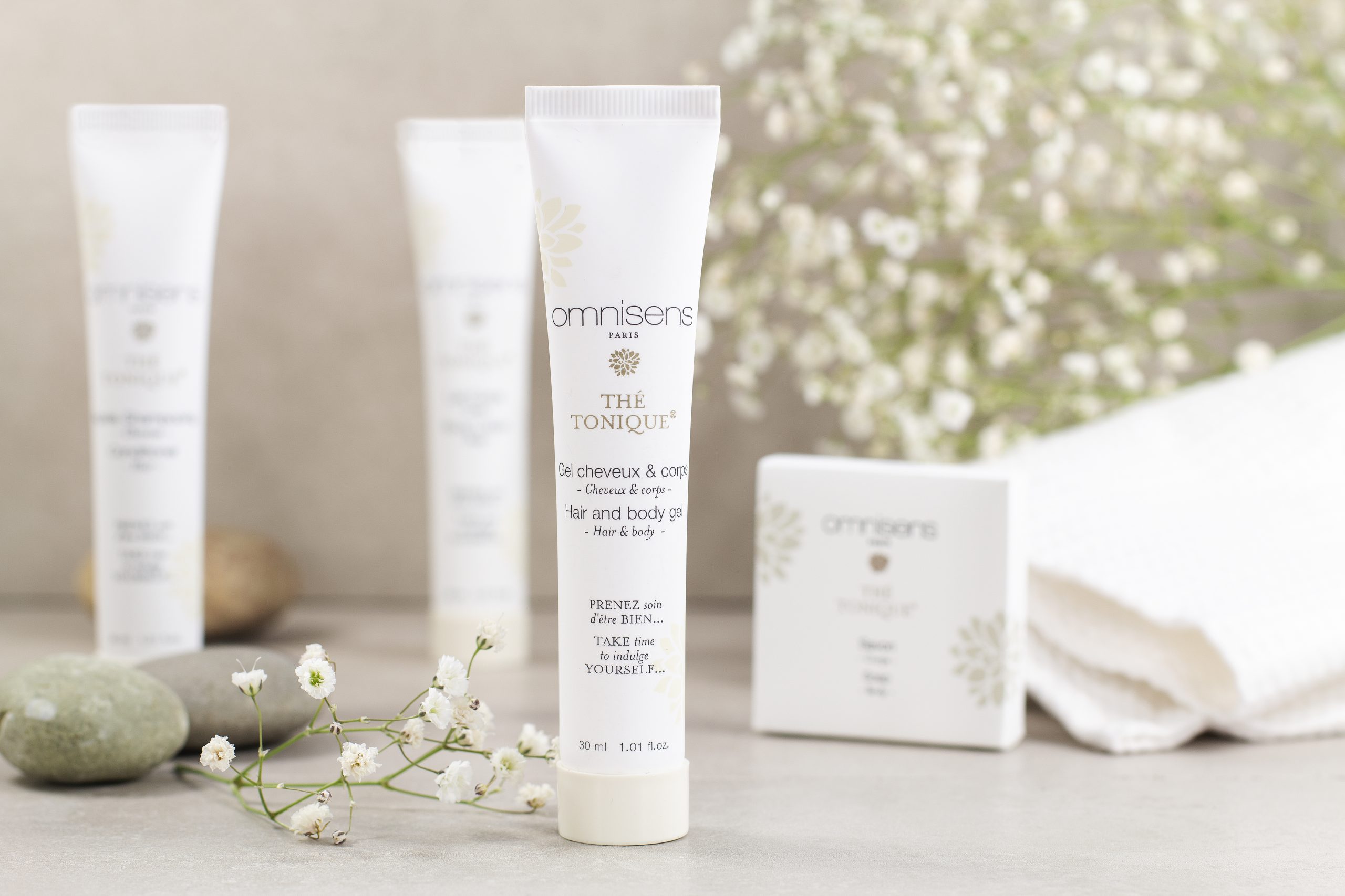 Travel-size Omnisens Paris “Thé Tonique” hair and body gel tube standing upright, surrounded by delicate white flowers and folded towels.