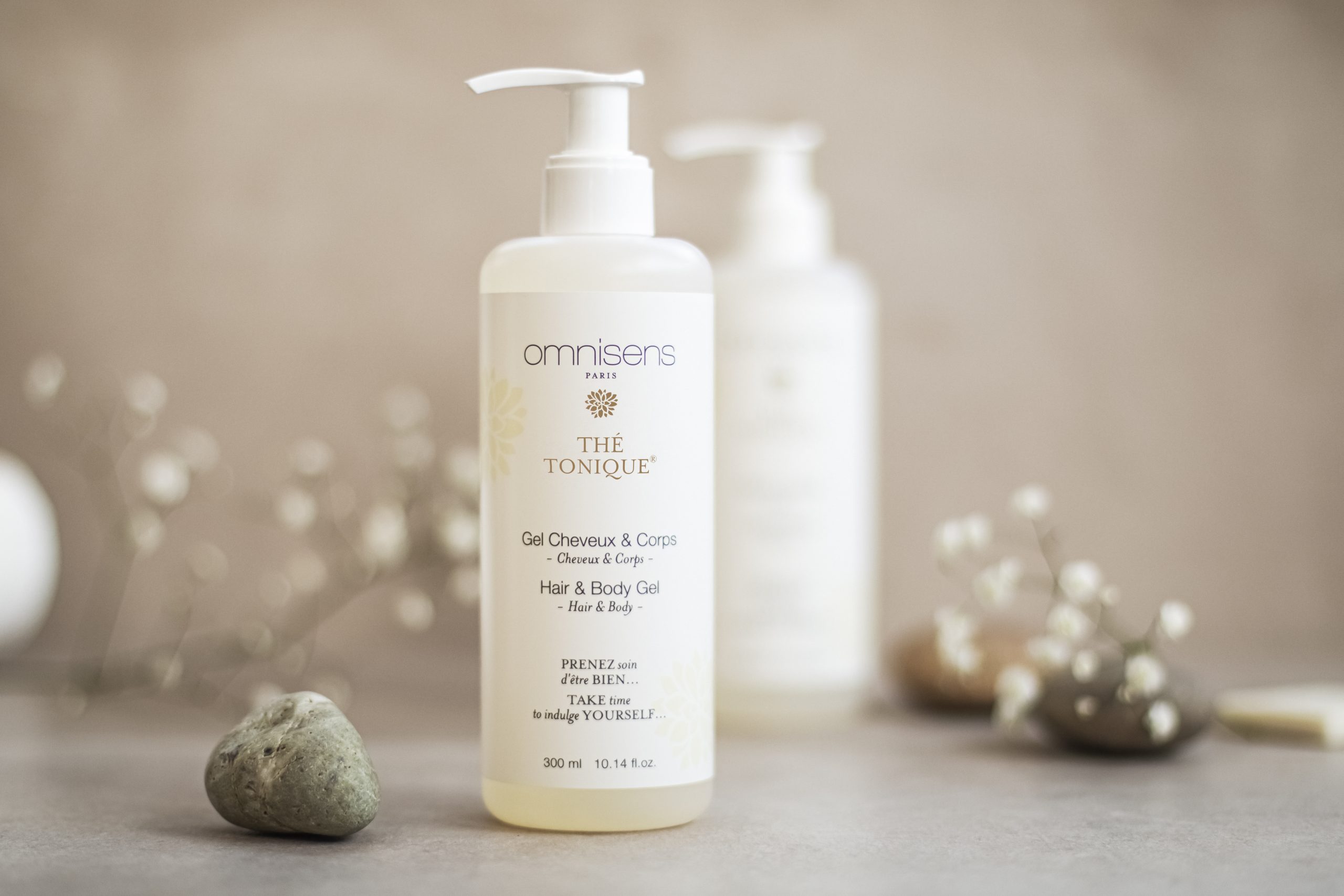 Omnisens Paris “Thé Tonique” hair and body gel bottle displayed on a neutral surface with stones and soft floral accents.