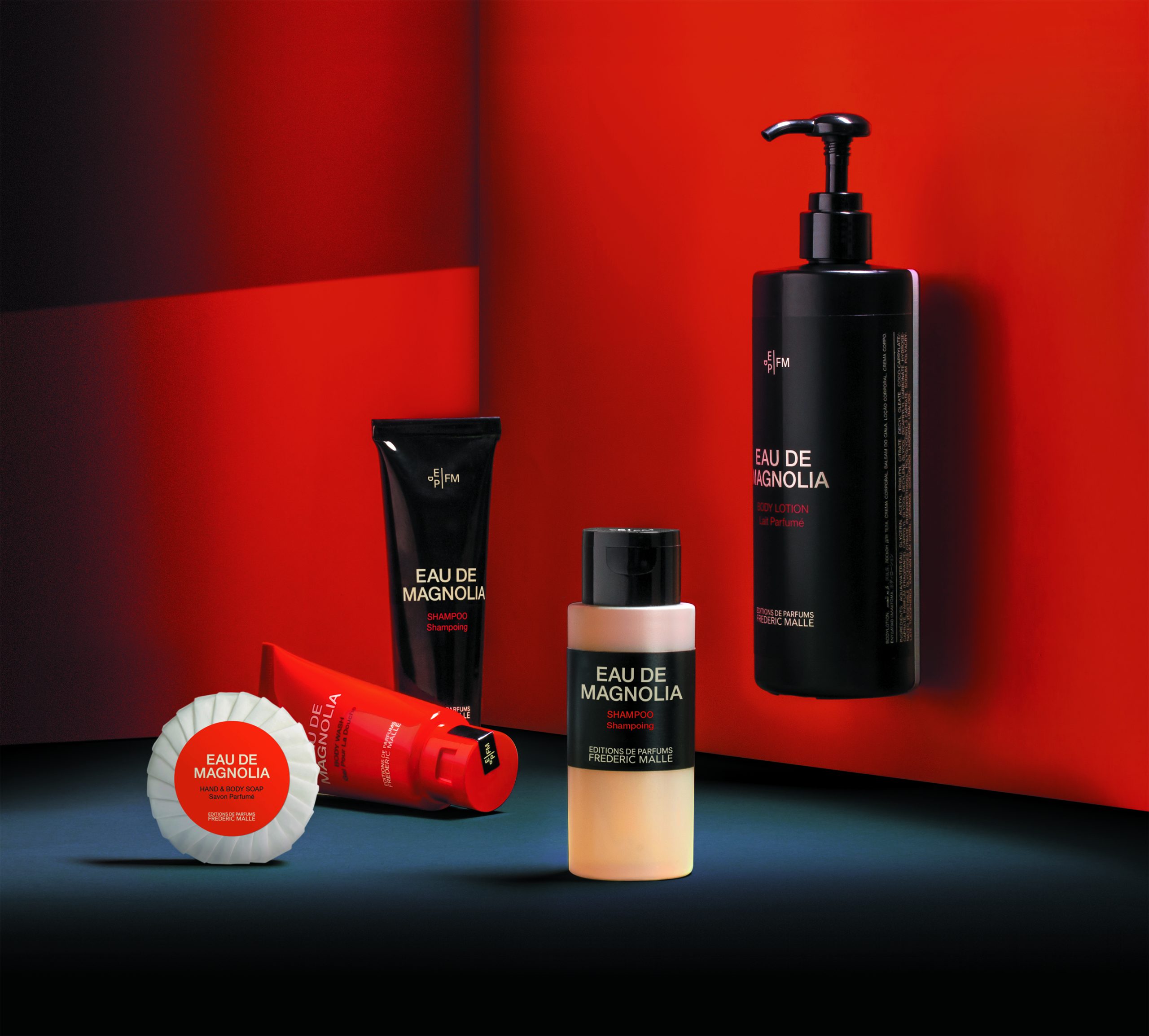 Editions de Parfums Frédéric Malle Eau de Magnolia product range displayed against a bold red background, including body lotion with pump, shampoo tube, body wash tube, and round wrapped soap, arranged with strong lighting and deep shadows.