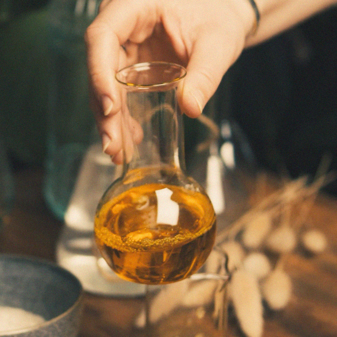 Hand holding a small glass bottle filled with golden liquid, with natural ingredients and a soft, warm-toned background suggesting a cosmetic or skincare formulation.