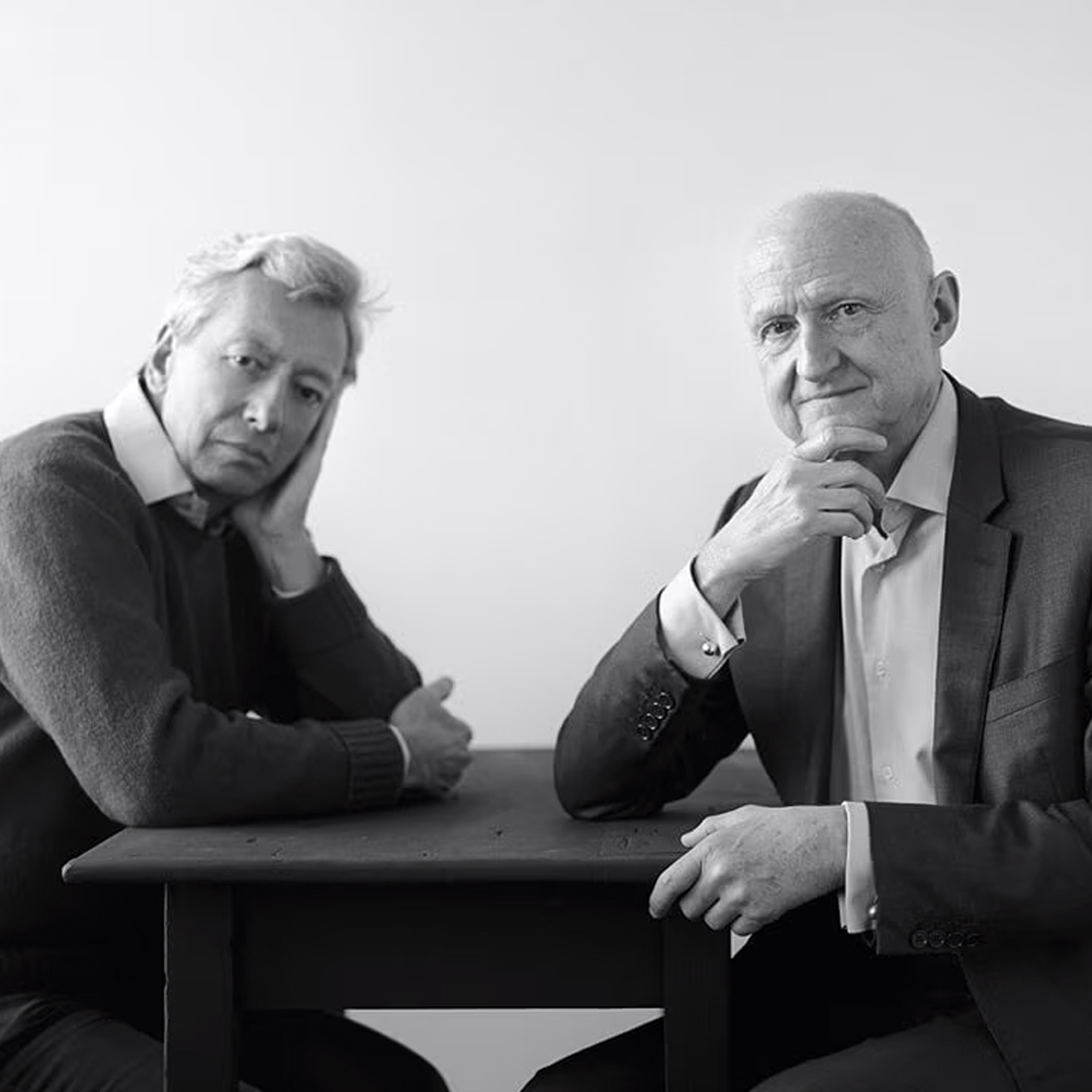 Black-and-white portrait of Frederic Malle and Dominique Ropion seated at a table, facing the camera in a minimalist studio setting with soft lighting and a neutral background.