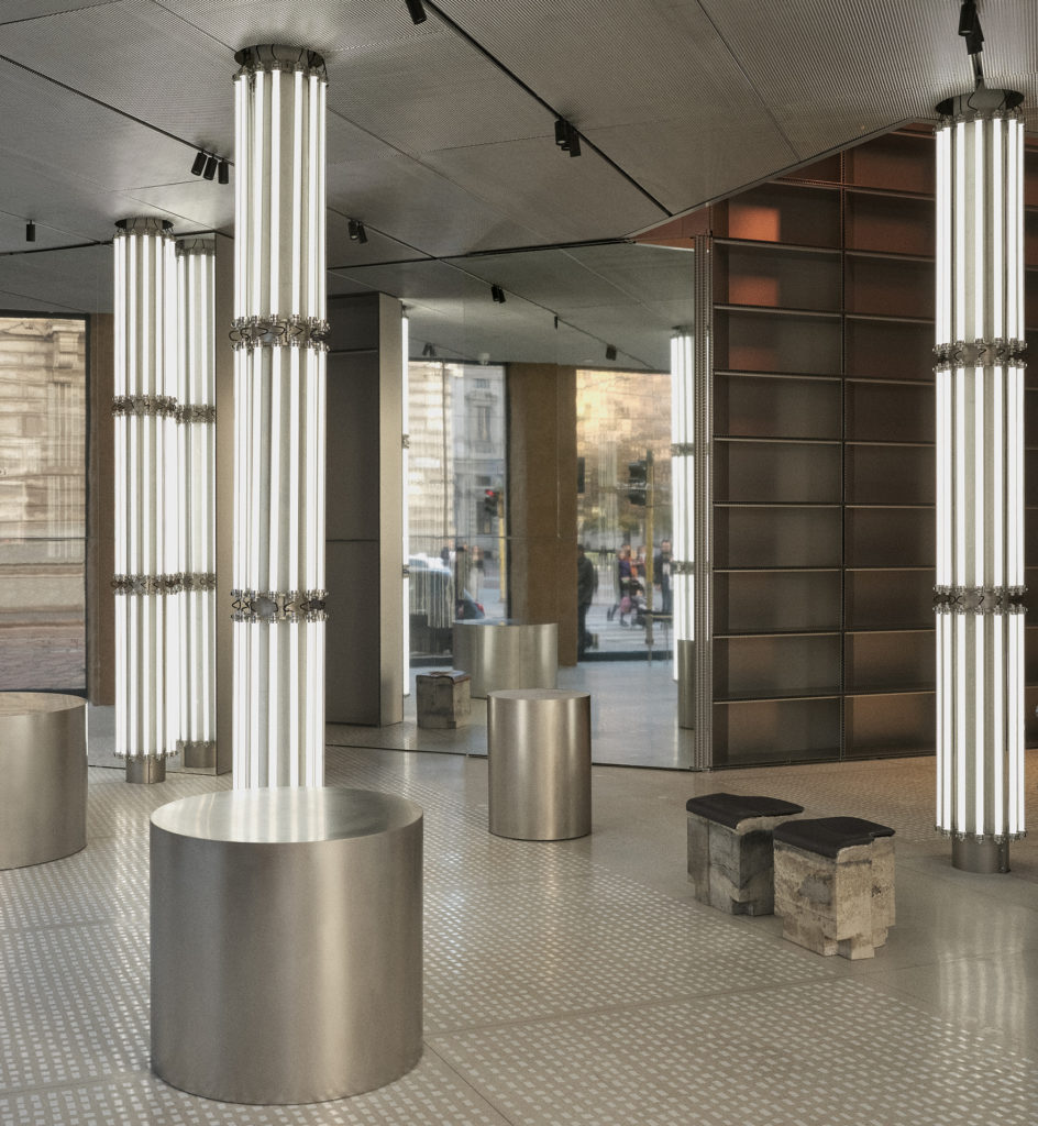 Modern retail or showroom interior with metallic cylindrical displays, tall vertical light fixtures, and reflective surfaces, creating a sleek and contemporary architectural space.