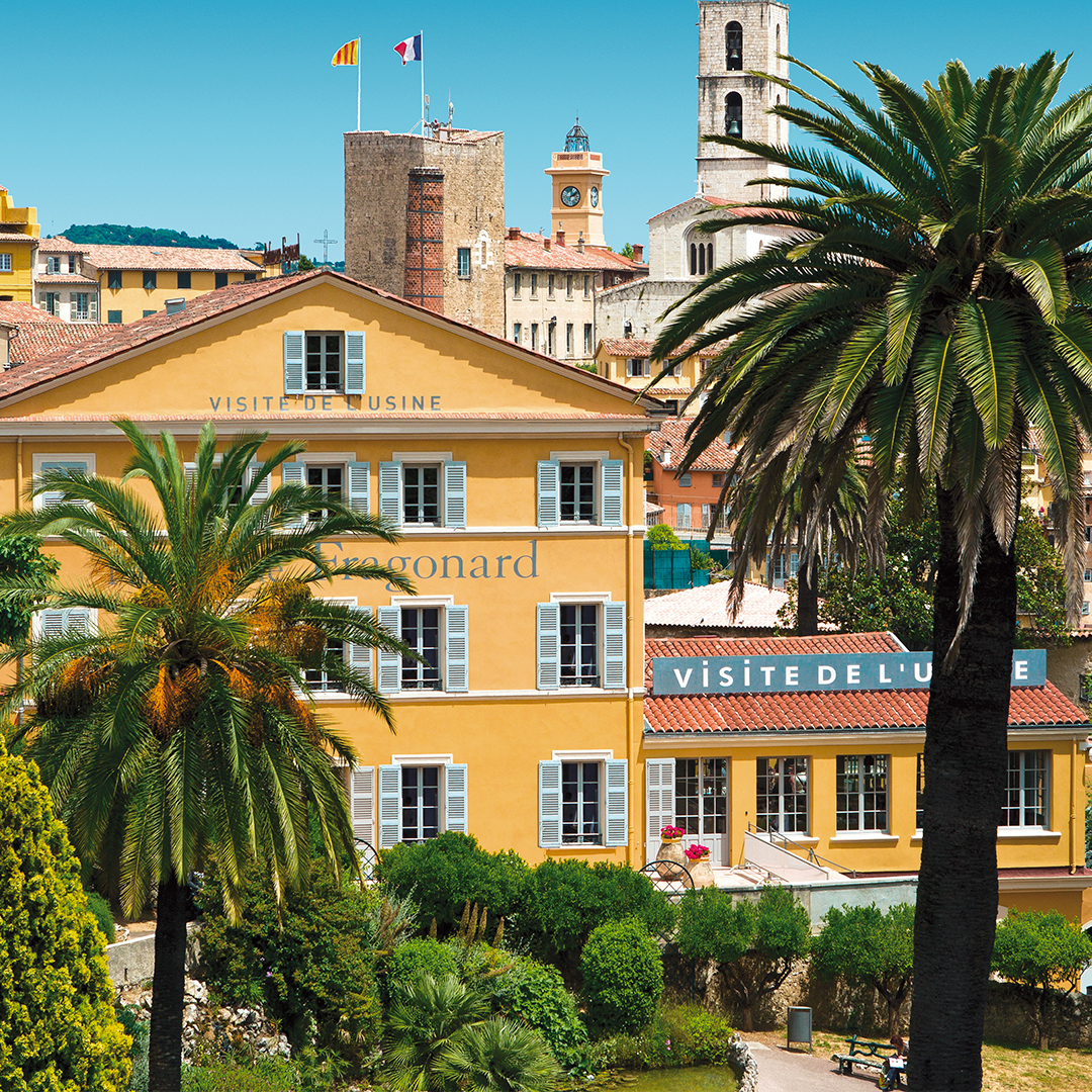 Fragonard factory building in a sunny Mediterranean setting, with yellow walls, green shutters, palm trees, and visible signage reading “VISITE DE L’USINE,” surrounded by historic architecture and a blue sky.