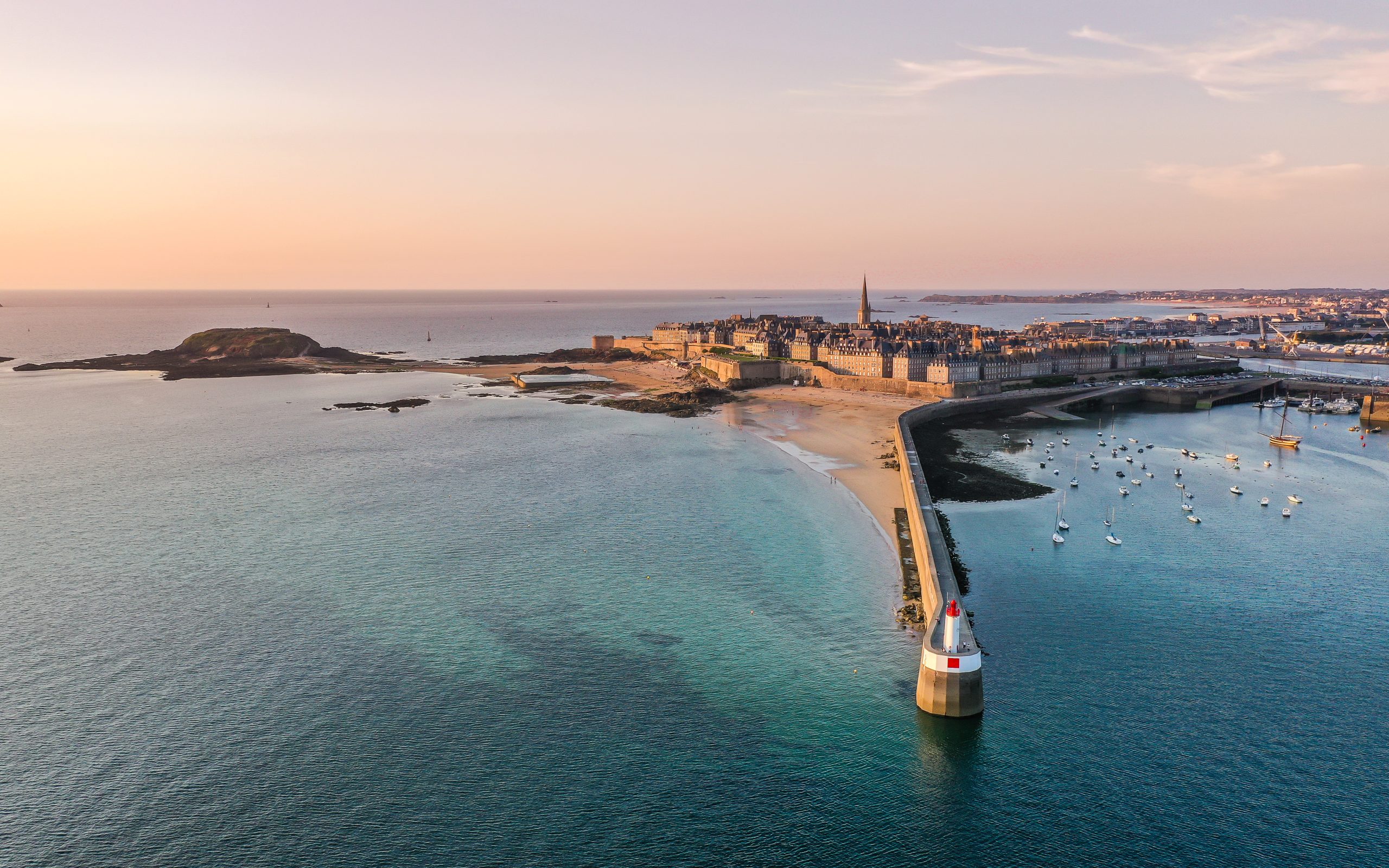 Vue aérienne du littoral de Saint-Malo avec les remparts, le port et le phare au coucher du soleil.