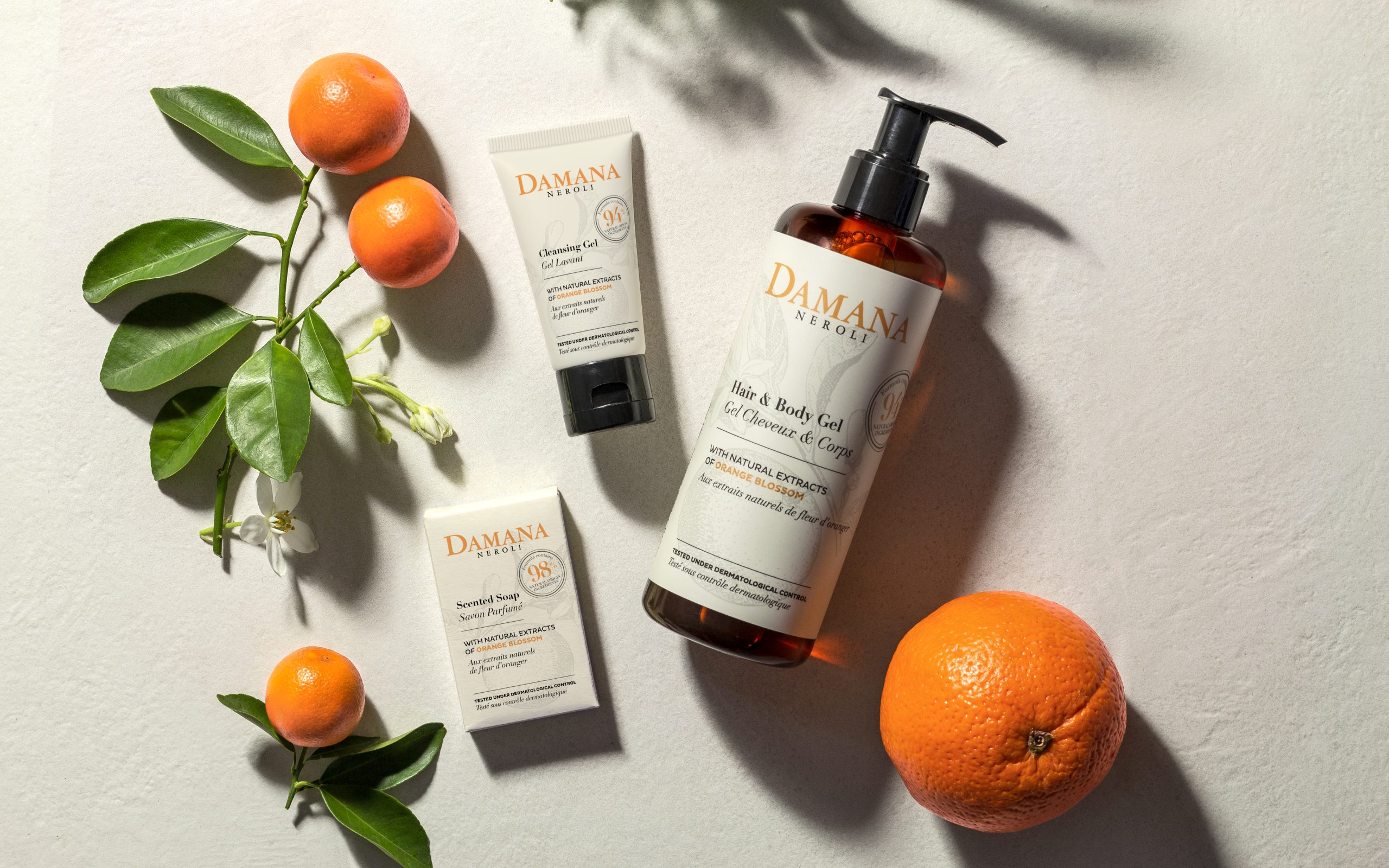 Damana Neroli skincare products displayed on a light surface alongside fresh oranges and green leaves, including a hair and body gel bottle, cleansing gel tube, and scented soap, highlighting natural orange blossom ingredients and a fresh, citrus-inspired aesthetic.