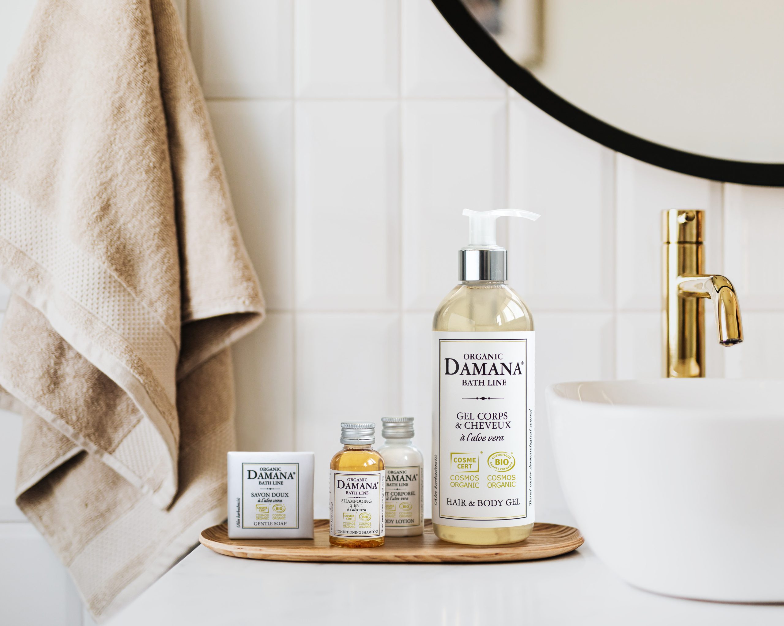 Organic Damana bath products arranged on a wooden tray beside a sink, including a large hair and body gel bottle, mini shampoo and lotion bottles, and a boxed soap, with soft towels and a modern bathroom setting in the background.