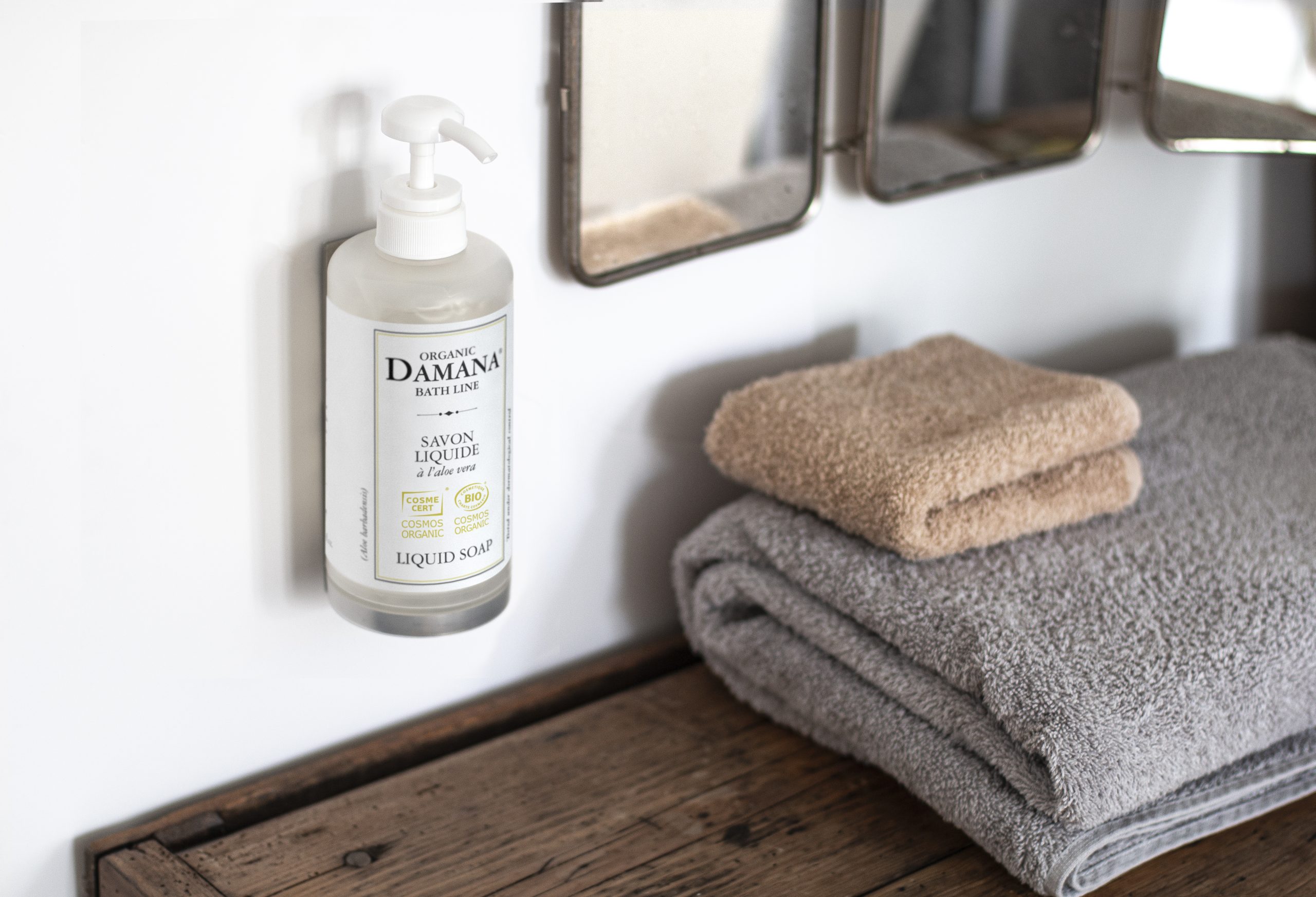 Wall-mounted bottle of Organic Damana liquid soap next to neatly stacked beige and gray towels, set in a clean bathroom environment with a mirror and wooden shelf.