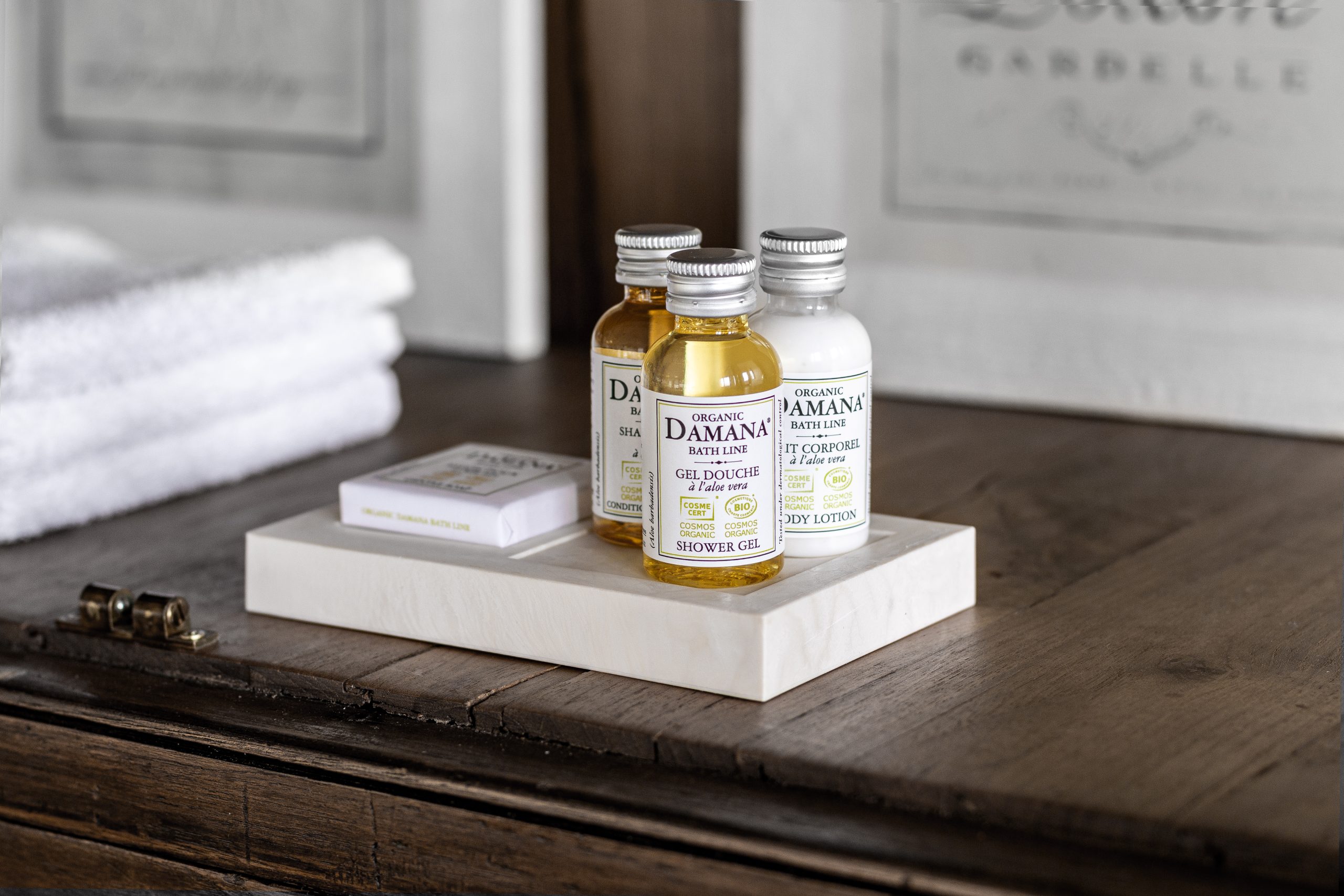 Organic Damana bath amenities displayed on a white tray on a wooden surface, including small bottles of shower gel, shampoo, and body lotion alongside a boxed soap, with folded towels in the background.