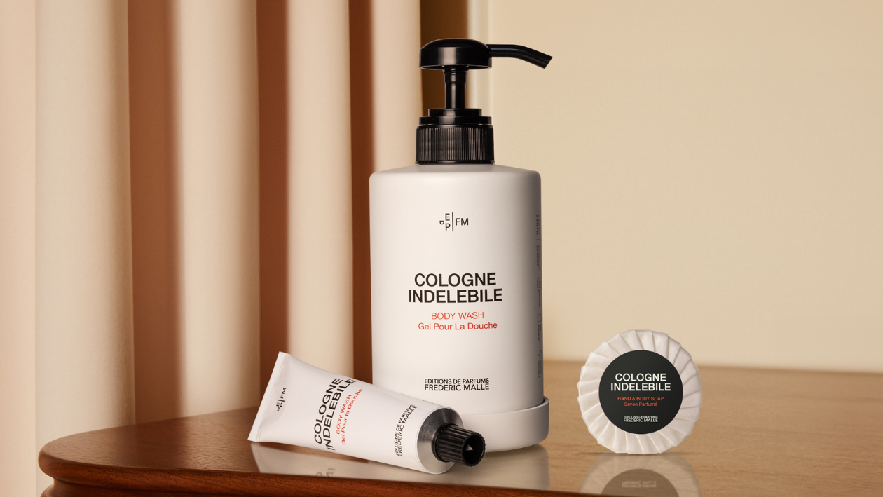 Cologne Indélébile body wash bottle with pump, bath foam tube, and wrapped soap displayed on a wooden surface, highlighted by warm lighting and a minimalist beige background.