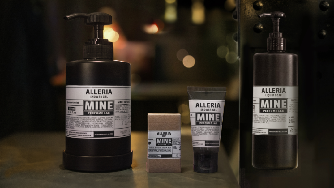 Alleria Mine Perfume Lab bath product lineup displayed in a dimly lit setting, including a large shower gel bottle, liquid soap pump, a small tube, and a boxed soap, with warm bokeh lights creating a cozy, refined atmosphere.