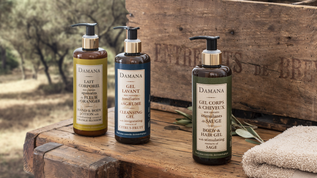 Three Damana cosmetic pump bottles displayed on a rustic wooden surface outdoors, including body lotion, cleansing gel, and body and hair gel, with natural elements and a folded towel nearby.