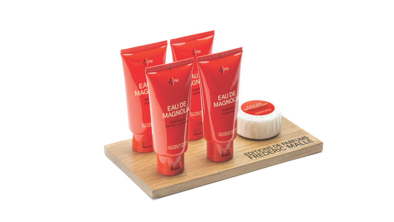 Editions de Parfums Frédéric Malle Eau de Magnolia products arranged on a wooden tray, featuring multiple red tubes (body wash and body lotion) alongside a wrapped round soap with a red label, set against a clean, minimalist background.