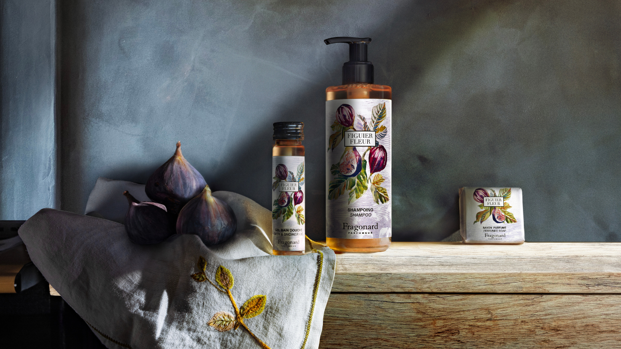 Fragonard Figuer Fleur bath products displayed on a wooden surface with fresh figs and fabric, including a shower gel bottle, shampoo pump, and soap bar, set against a textured background with soft, natural lighting.