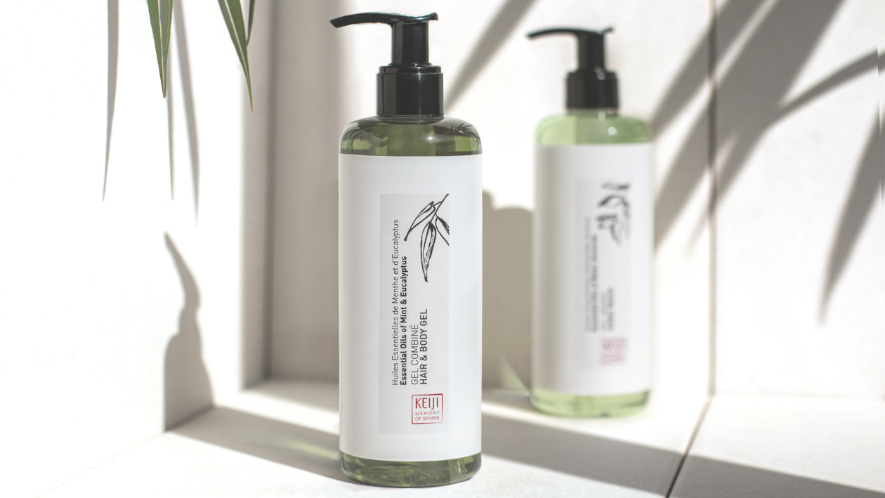 Keiji Memory of Senses body lotion bottle in the foreground with two matching green-toned bottles softly blurred in the background, set against a bright minimalist backdrop with soft natural shadows and plant leaves.