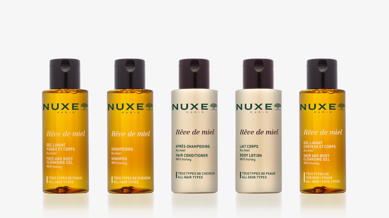 Gama de productos «Rêve de miel» de NUXE Paris, presentados sobre un fondo claro, que incluye gel limpiador facial y corporal, champú, acondicionador, loción corporal y gel limpiador para el cabello y el cuerpo en envases de color ámbar y beige.