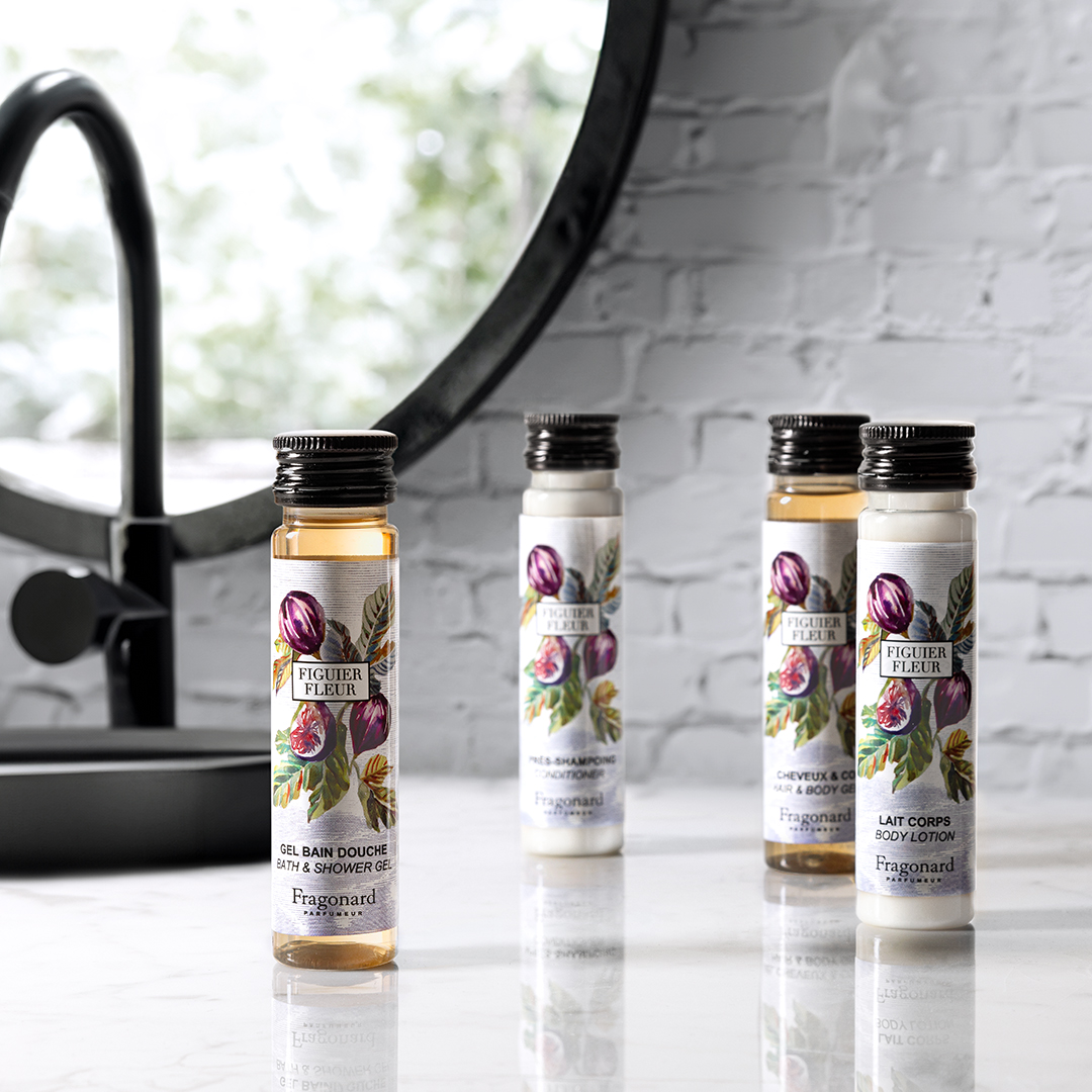 Fragonard Figuer Fleur travel-size bath products arranged on a marble surface near a sink and round mirror, featuring small bottles of shower gel, shampoo, conditioner, and body lotion with illustrated fig labels.
