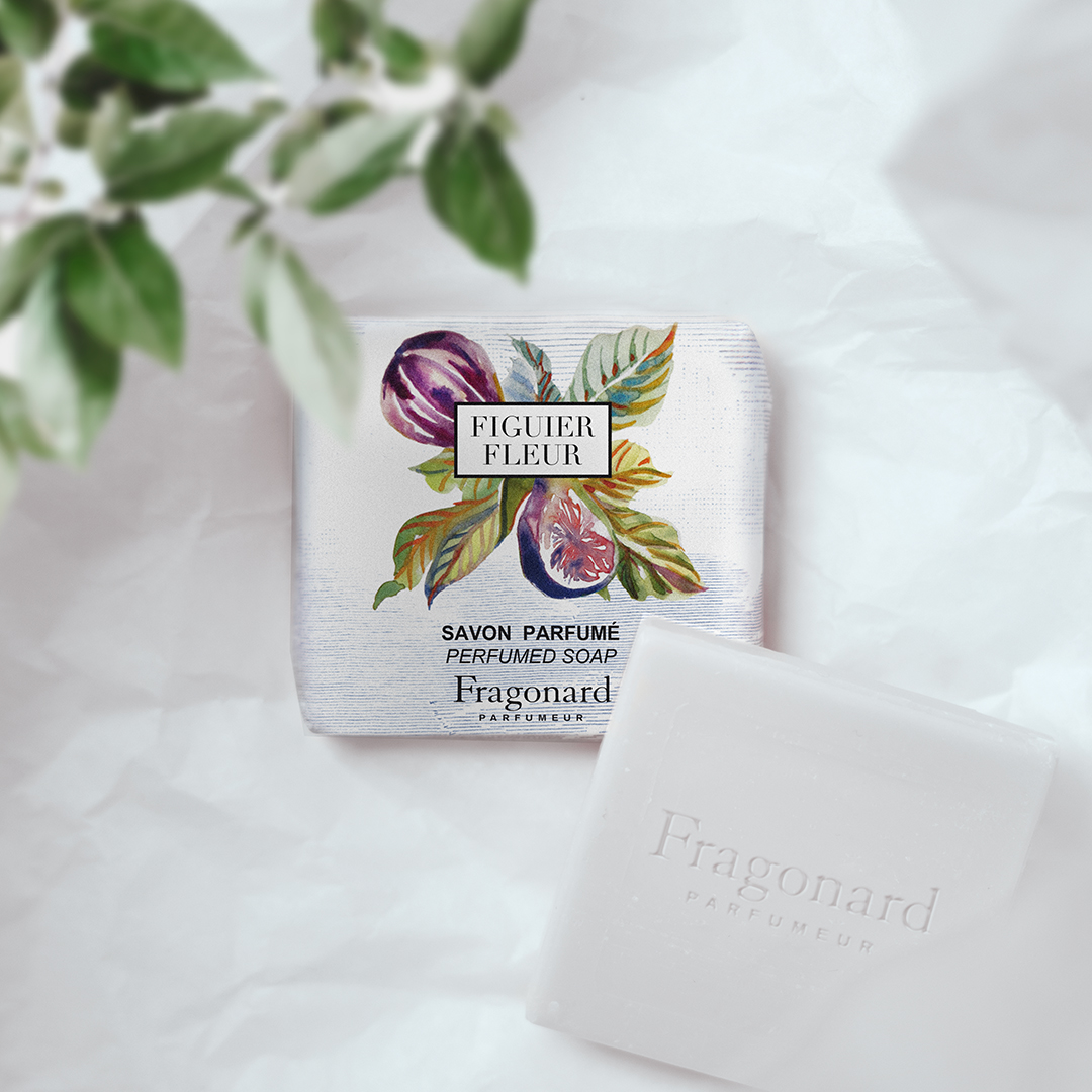 Fragonard Figuer Fleur perfumed soap in its illustrated packaging placed on a soft white surface, alongside the unwrapped white soap bar embossed with the Fragonard name, with leaves softly blurred in the foreground.