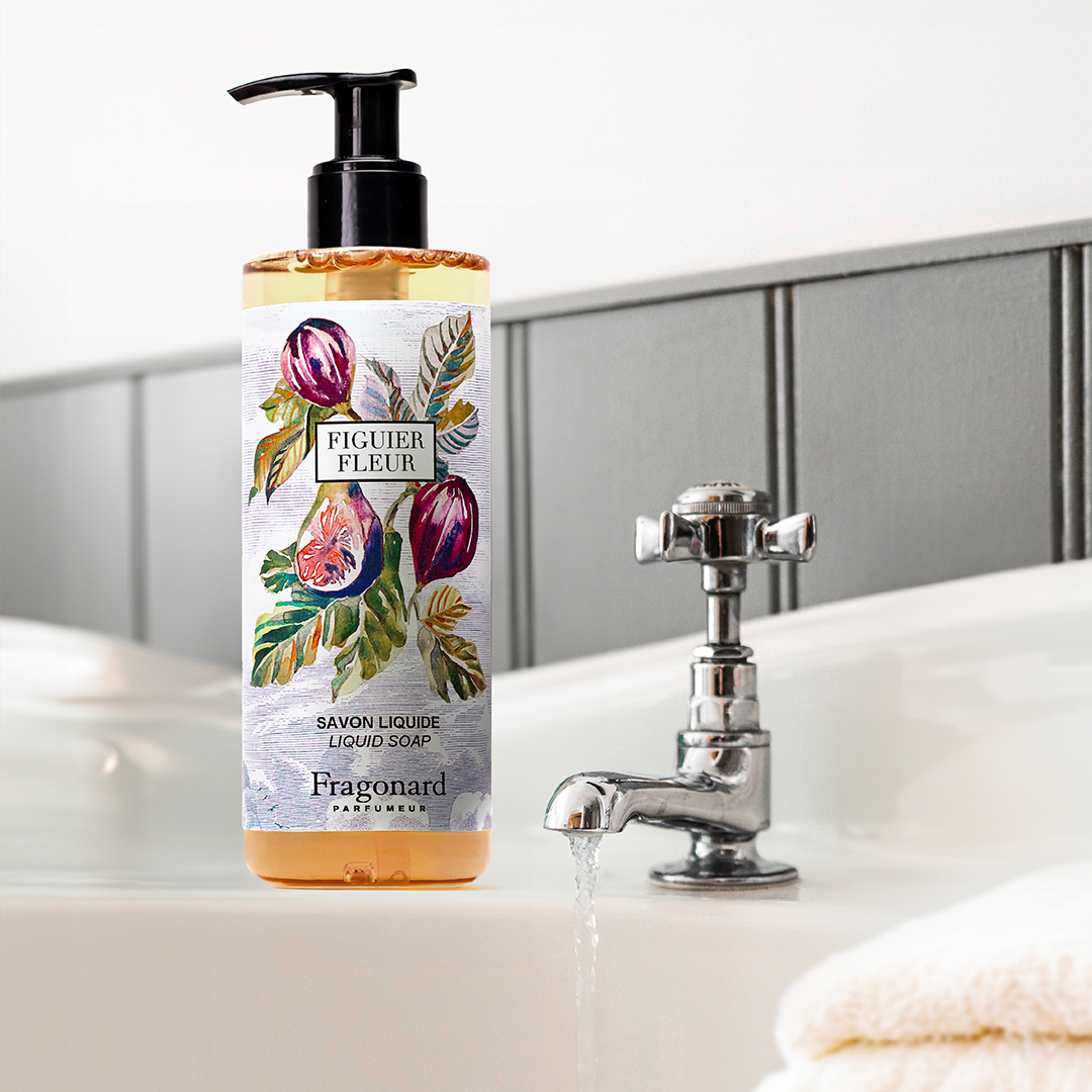 Fragonard Figuer Fleur liquid soap bottle positioned next to a white sink and chrome faucet, with water running and a folded towel nearby in a clean, minimalist bathroom setting.