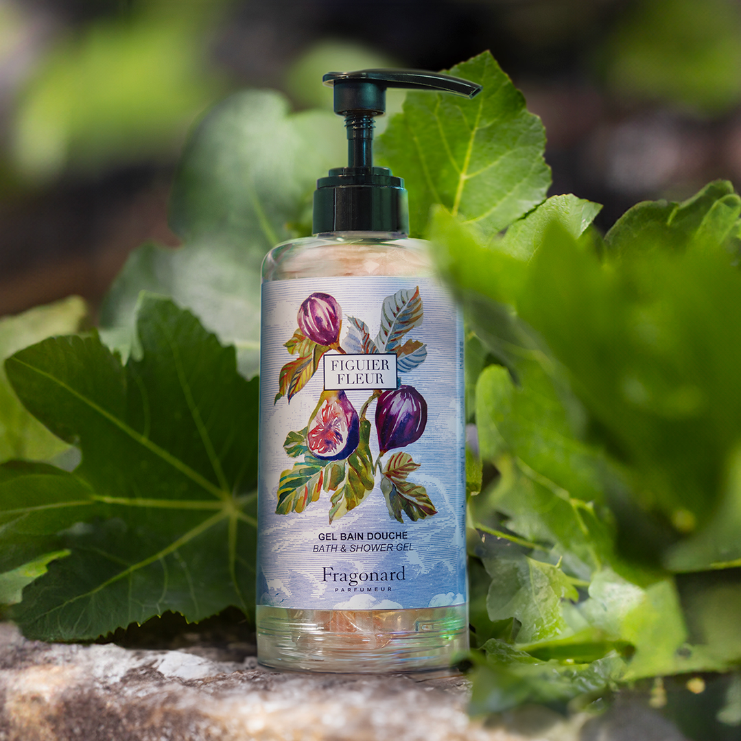 Fragonard Figuer Fleur bath and shower gel bottle with a black pump placed outdoors among large green leaves, with natural light highlighting the botanical fig illustration on the label.