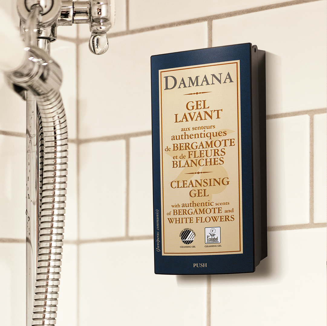 Wall-mounted Damana cleansing gel dispenser in a tiled shower, with a metal shower hose beside it and visible text highlighting bergamot and white flower scents.