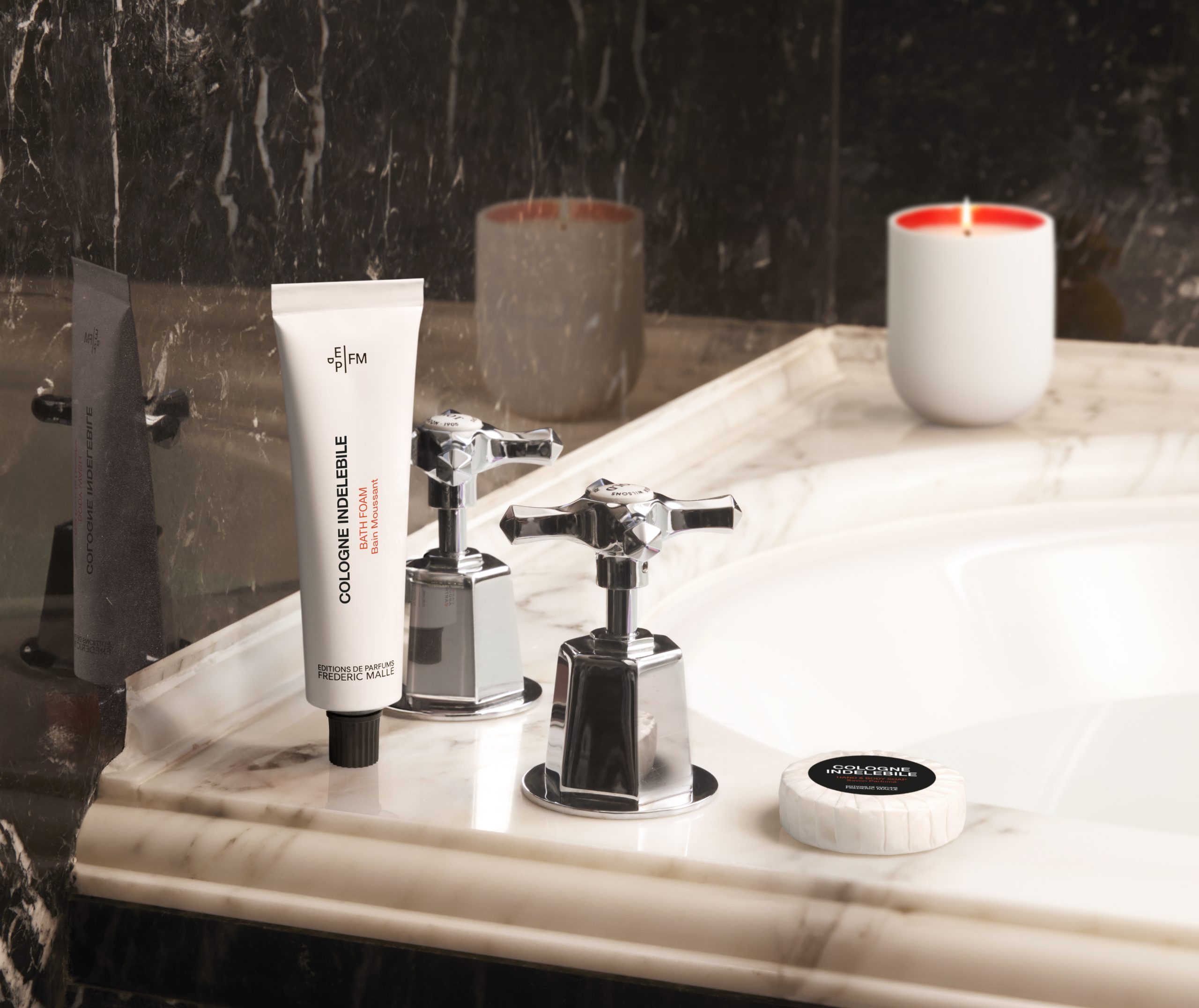 Cologne Indélébile bath foam tube by Editions de Parfums Frédéric Malle placed on a marble sink next to chrome faucet handles, with a soap bar and candle in a refined bathroom setting.