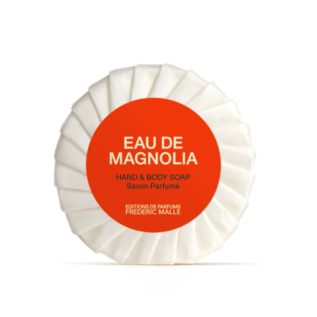 Round wrapped Eau de Magnolia hand and body soap by Editions de Parfums Frédéric Malle with a white pleated wrapper and a bright red circular label centered on a plain light background.