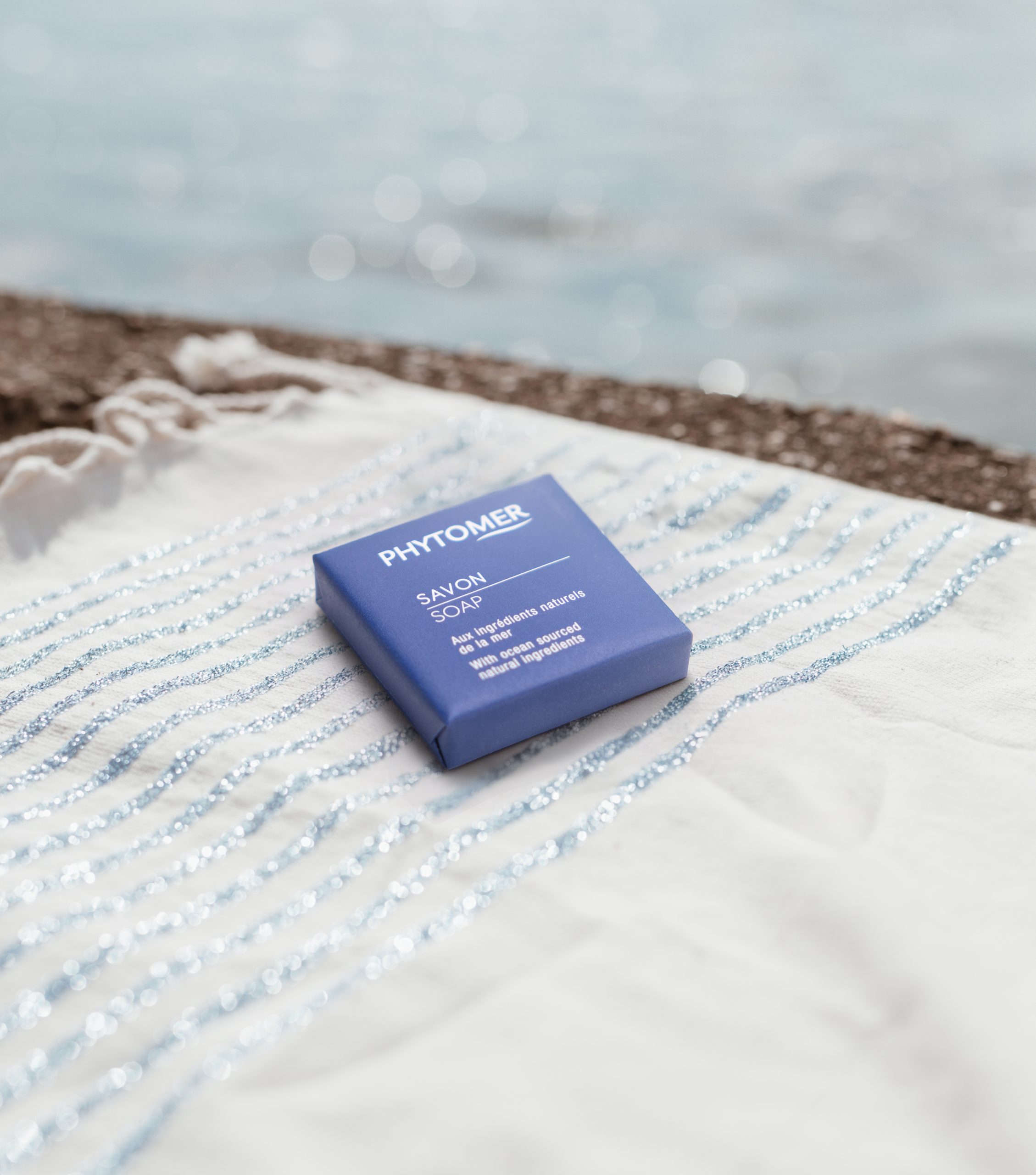 Phytomer savon emballé posé sur une serviette de plage rayée au bord de la mer.