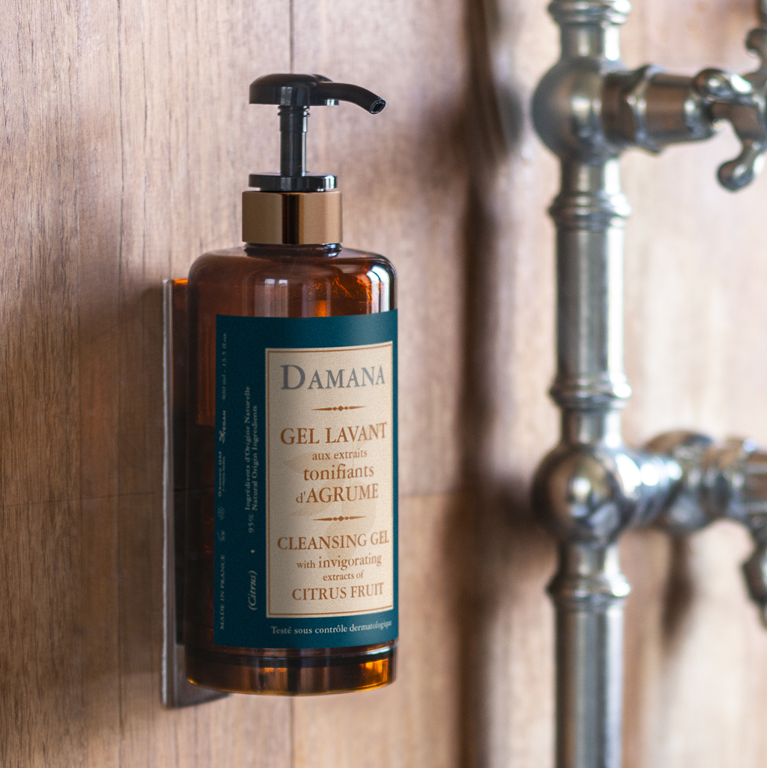Damana cleansing gel bottle with a pump dispenser mounted on a wooden wall, next to metal pipes, showcasing citrus extract ingredients and a rustic bathroom setting.