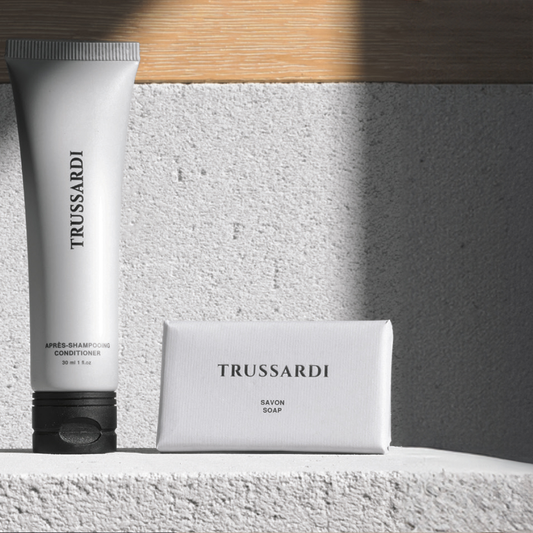 Trussardi conditioner tube standing upright next to a wrapped soap bar on a textured surface, illuminated by soft natural light that casts warm shadows and enhances the clean, minimal design.