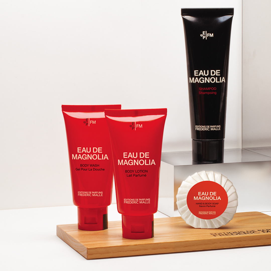 Eau de Magnolia bath products by Editions de Parfums Frédéric Malle arranged on a wooden tray, featuring two red tubes (body wash and body lotion), a black shampoo tube, and a wrapped soap, set in a clean, minimalist space.
