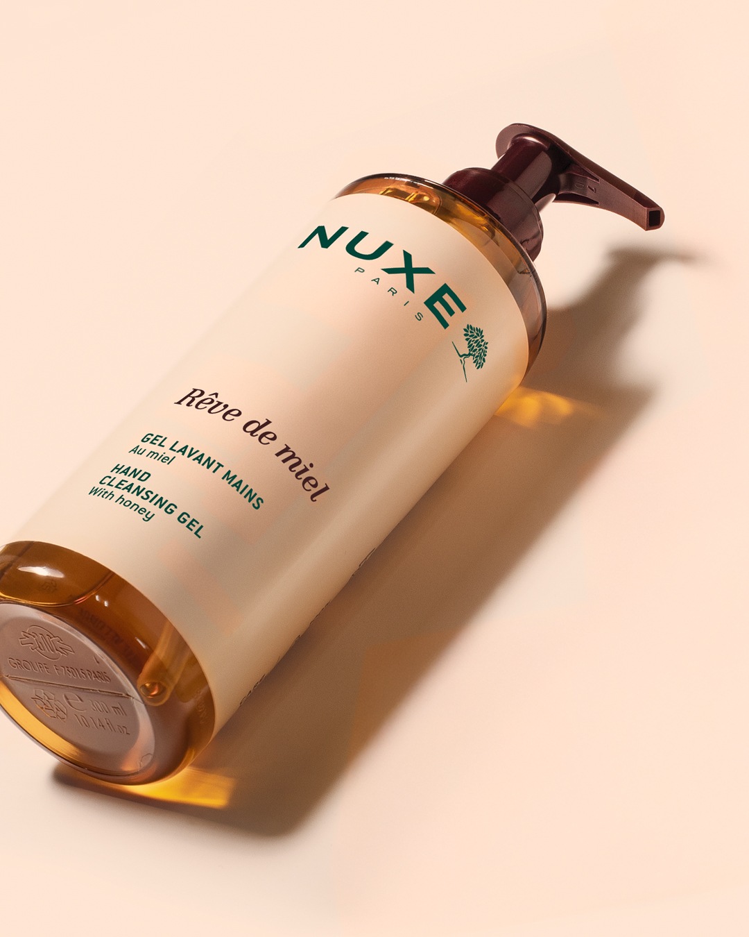 Gel limpiador de manos NUXE Rêve de miel en un botellas con dosificador, inclinado sobre un fondo beige, con el líquido ámbar a la vista y una iluminación suave que crea sombras elegantes.