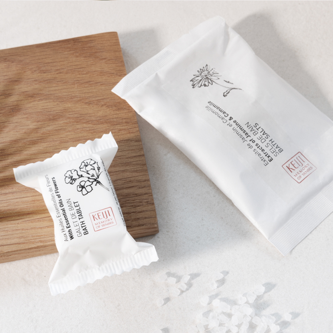 Keiji Memory of Senses bath products displayed on a light surface, featuring individually wrapped bath tablets and bath salts in white packaging next to a wooden tray, with scattered salt crystals adding a clean, spa-inspired feel.