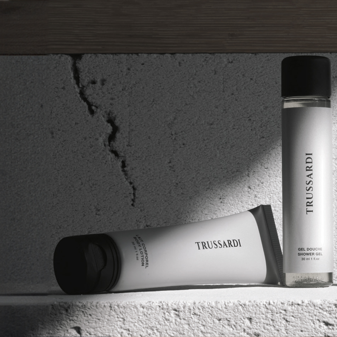 Trussardi body lotion tube and shower gel bottle displayed on a textured concrete shelf, with dramatic lighting creating strong shadows and highlighting the minimalist black-and-white packaging.